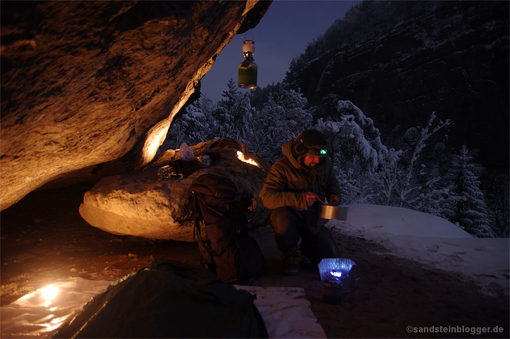 Mann mit Topf und Kocher unter Felsen, verschneiter Wald, Nacht