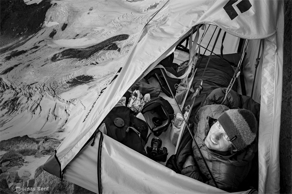 Mann imSchlafsack im Portaledge, neben ihm Kameras, Objektive und andere Fotoausrüstung
