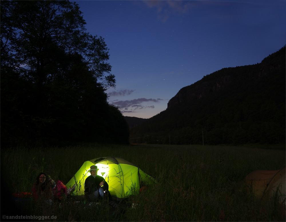 Von innen beleuchtetes Zelt, davor zwei Frauen und ein Mann, Nachthimmel, schemenhafte Felsen