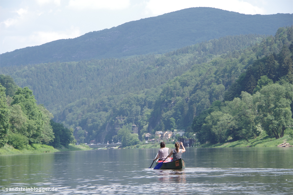 Zwei Frauen paddeln im Kanu den Fluss hinunter, am Ufer eine runde Bergkuppe