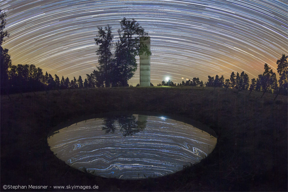 Metallplatte, in der sich der Sternenhimmel spiegelt, dahinter Aussichtsturm