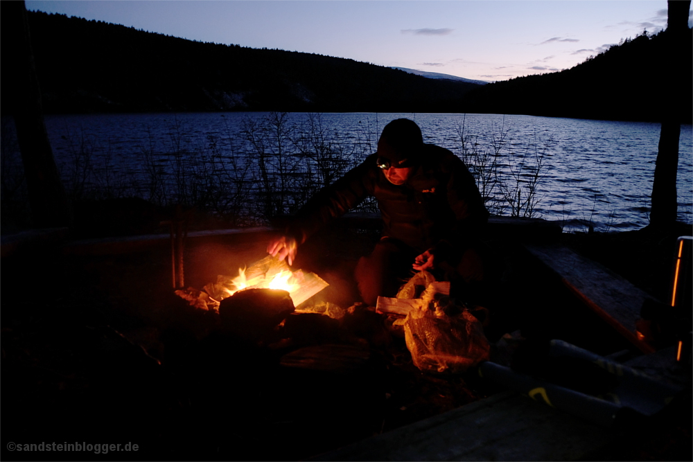 Mann am Lagerfeuer, dahinter ein Fluss in der Abenddämmerung