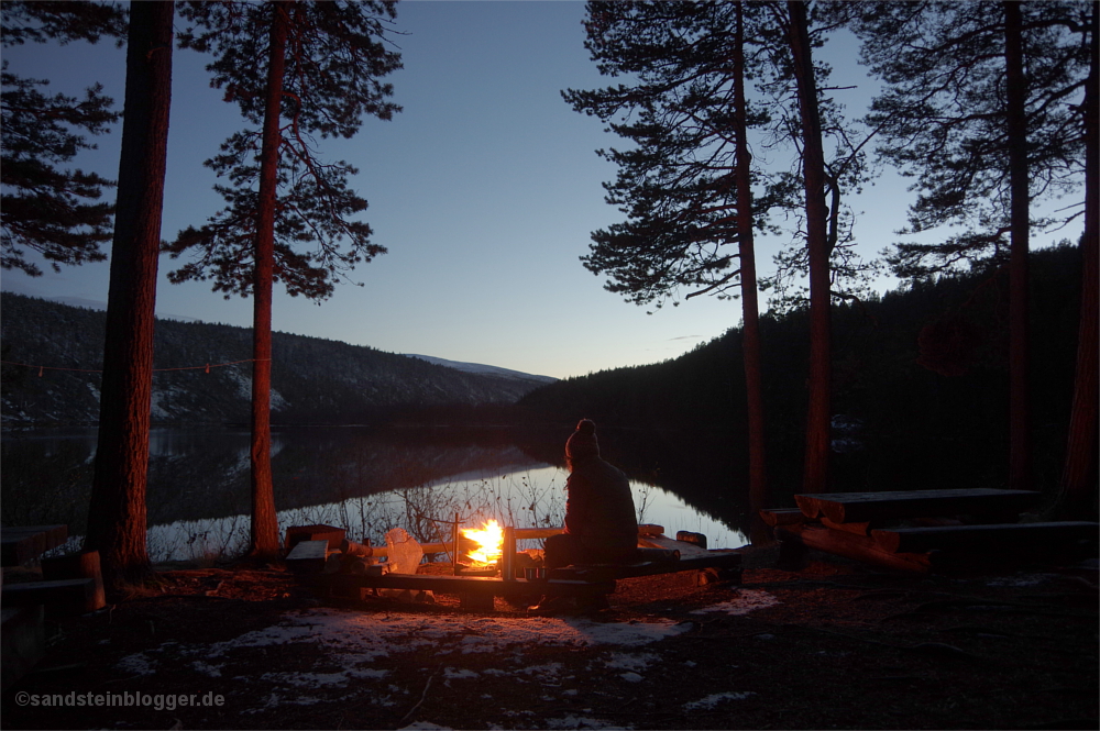 Frau am Lagerfeuer, dahinter Fluss in der Abenddämmerung