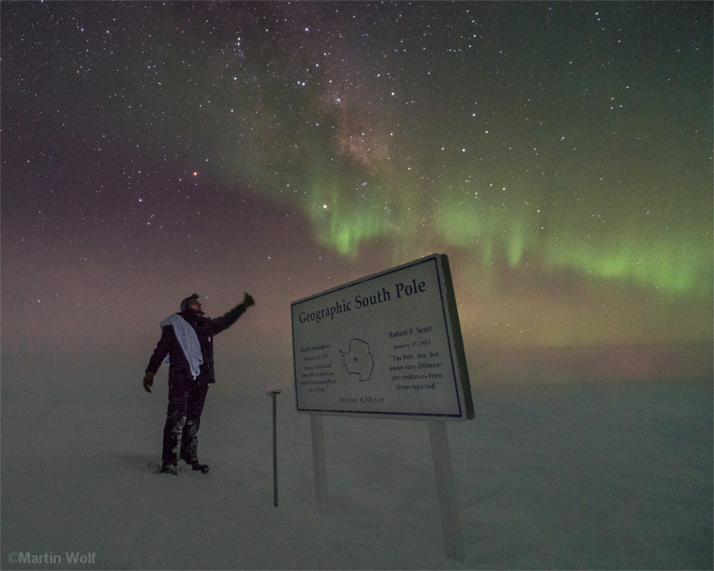 Mann steht am Südpol vor einer Gedenktafel, die an die Polarforscher Roald Amundsen und Robert Falcon Scott erinnert. Über seinem Kopf Polarlichter.