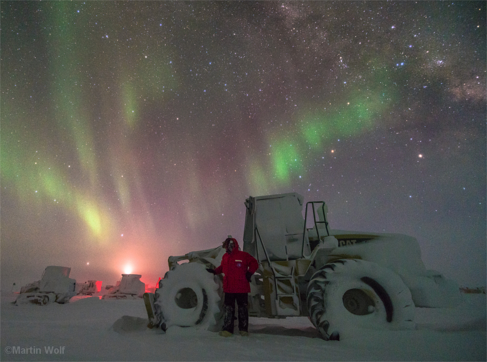 Mann vor verschneiter Schneefräse, darüber die Polarlichter