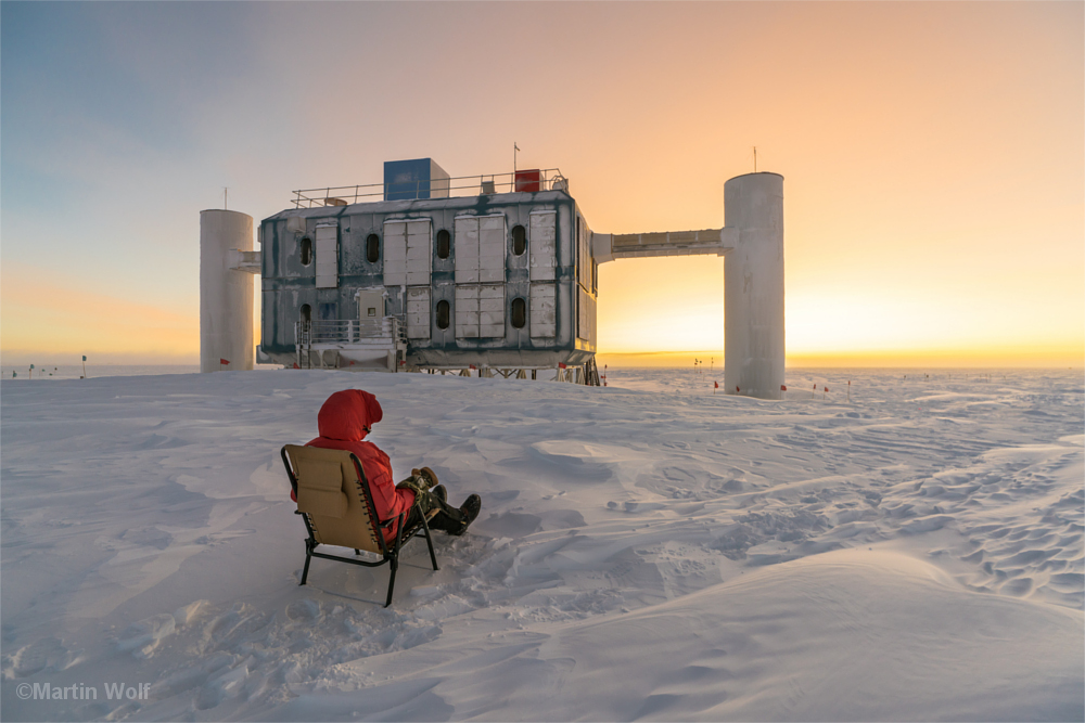 Mann in dicker Daunenjacke sitzt in einem Liegestuhl mitten auf dem Eisfeld am Südpol - vor sich einen Teil der Forschungsgebäude der Amundsen-Scott-Station.