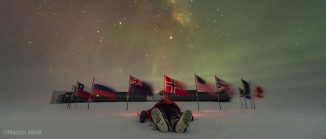 Mann liegt im Schnee. Dahinter ein Kreis von Flaggen, die den Südpol markieren. In der Ferne die Gebäude der Amundsen-Scott-Forschungsstation. Am nächtlichen Himmel tanzen Polarlichter.