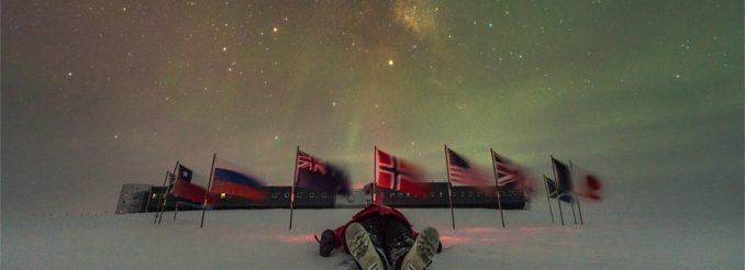 Mann liegt im Schnee. Dahinter ein Kreis von Flaggen, die den Südpol markieren. In der Ferne die Gebäude der Amundsen-Scott-Forschungsstation. Am nächtlichen Himmel tanzen Polarlichter.