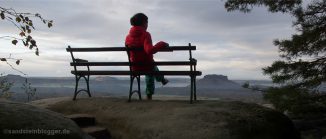 Eine Frau sitzt auf einer Bank. Von dort geht der Blick weit über die Tafelberge der Sächsischen Schweiz.