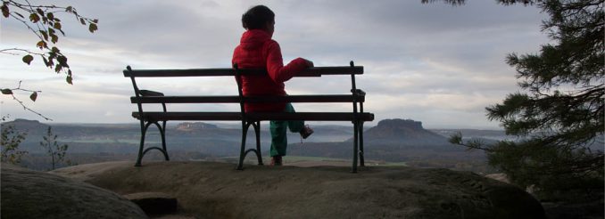 Eine Frau sitzt auf einer Bank. Von dort geht der Blick weit über die Tafelberge der Sächsischen Schweiz.