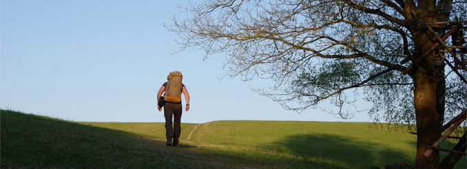 Mann mit Rucksack wandert über einen Wiesenhügel