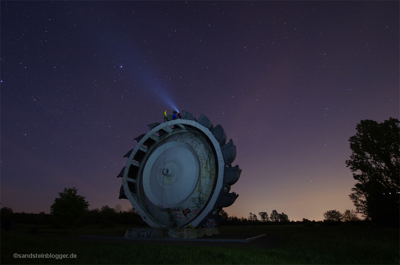 Schaufelrad eines Braunkohlebaggers, von seinem Scheitelpunkt geht ein Lichtstrahl zum Himmel