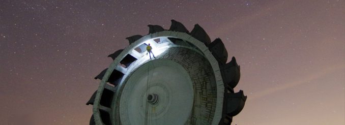 Schaufelrad eines Braunkohlebaggers vor dem Nachthimmel, ein Kletterer seilt sich daran ab.