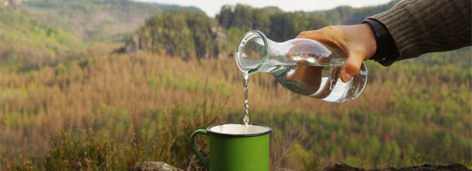 Felslandschaft, davor gießt jemand Wasser aus einer Glaskaraffe in einen Trinkbecher