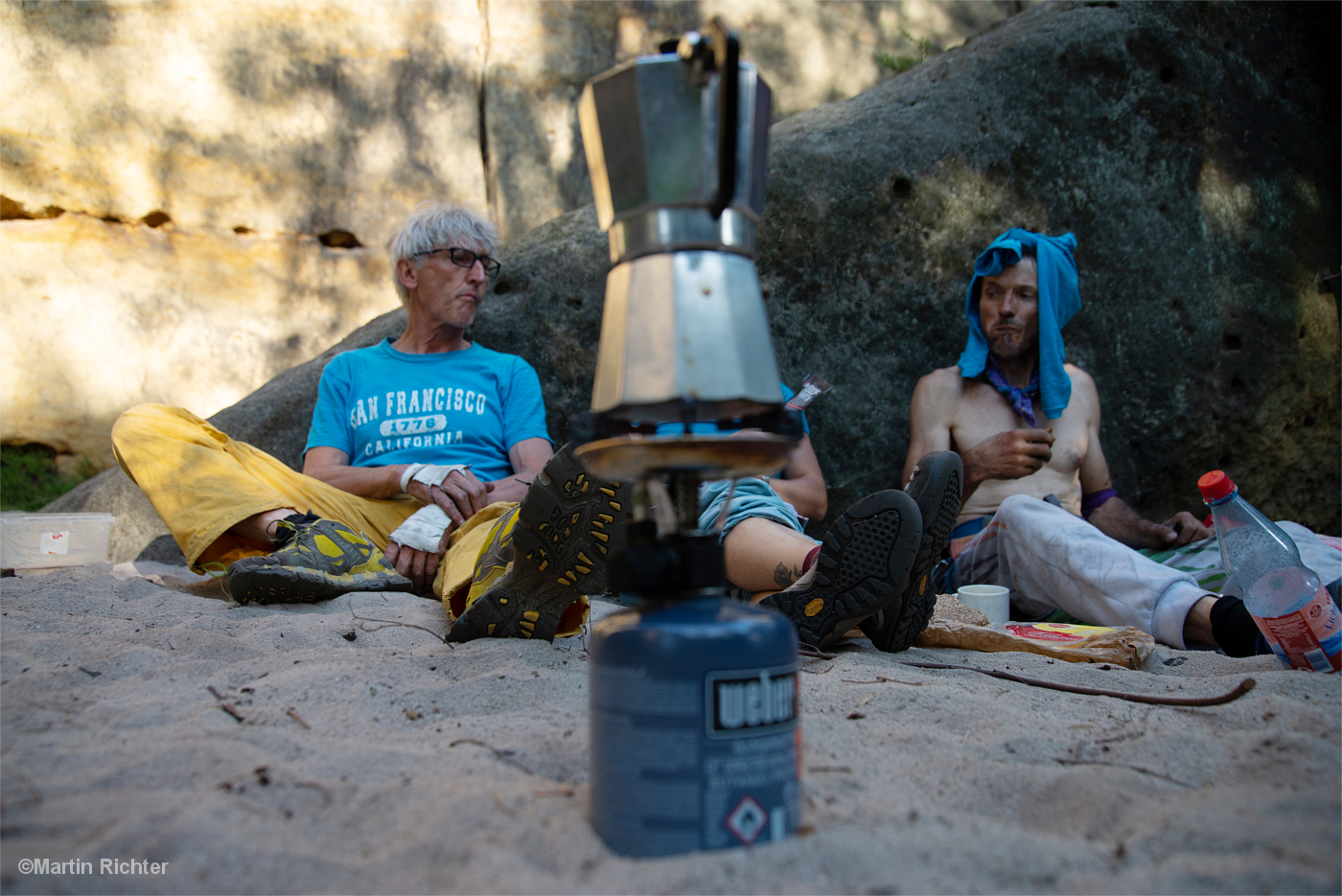 Zwei Kletterer sitzen vor einem Felsen im Sand, im Vordergrund eine Espressomaschine